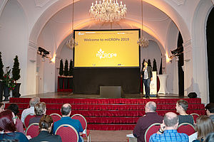 Angela Sessitsch vor dem Publikum auf der Bühne, hinter ihr eine große Leinwand mit dem Willkommens-Schriftzug der miCROPe Konferenz