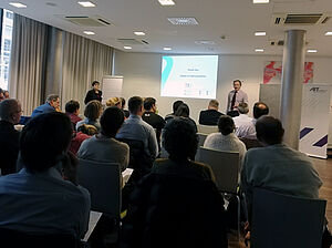 Meeting room, a guy justs presents in front of the audience, in the background his presentation is running next to the ait roll-up