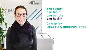Clara Pogner steht vor dem MobileAirClenaer, daneben steht auf Englisch: Eine Expertin, Ein Thema, Eine Minute, Eine Gesundheit, Zentrum für Gesundheit und Bioresourcen