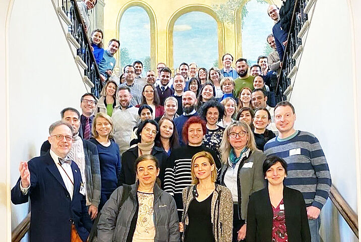 ine große Gruppe von Menschen steht für ein gemeinsames Foto auf einer breiten Treppe in einem hellen Gebäude mit bemaltem Gewölbe.