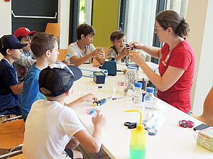 [Translate to English:] Children sit around a table and are taught by a scientist how to isolate samples