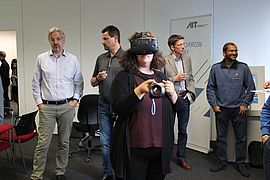 Das Bild zeigt eine Frau mit VR-Brille. Im Hintegrund unterhalten sich Menschen.