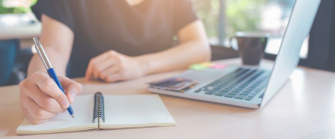 Person schreibt auf Block, Laptop steht daneben