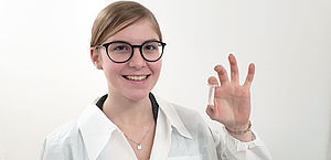 EIne junge Frau in weißem Labormantel hält lächelnd einen kleinen Probebehälter hoch in dem klein der weiße milchige Faden der extrahierten DNA zu sehen ist.