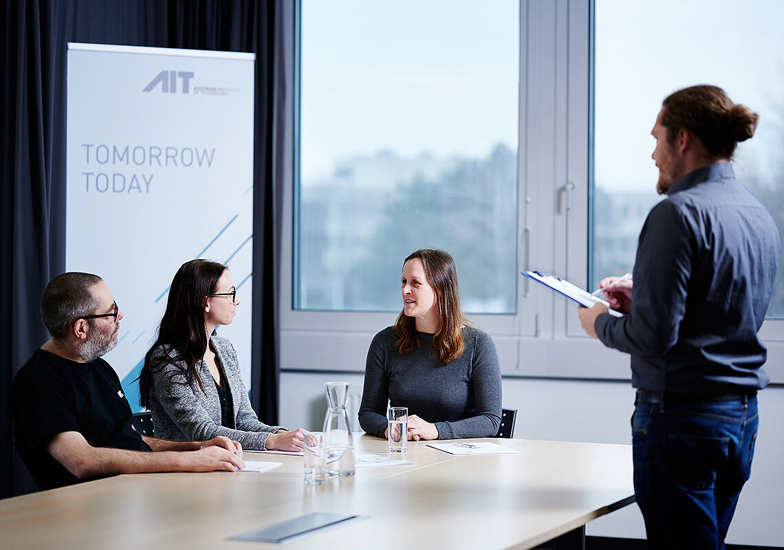 3 Personen sitzen am Tisch und unterhalten sich. Ein Mann beobachtet und macht Notizen.