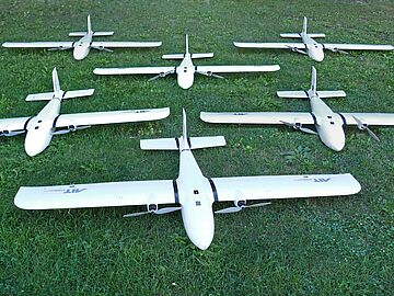 Several unmanned aerial vehicles equipped with sensors and the AIT logo lie on the meadow in front of the AIT Aerial Systems Lab