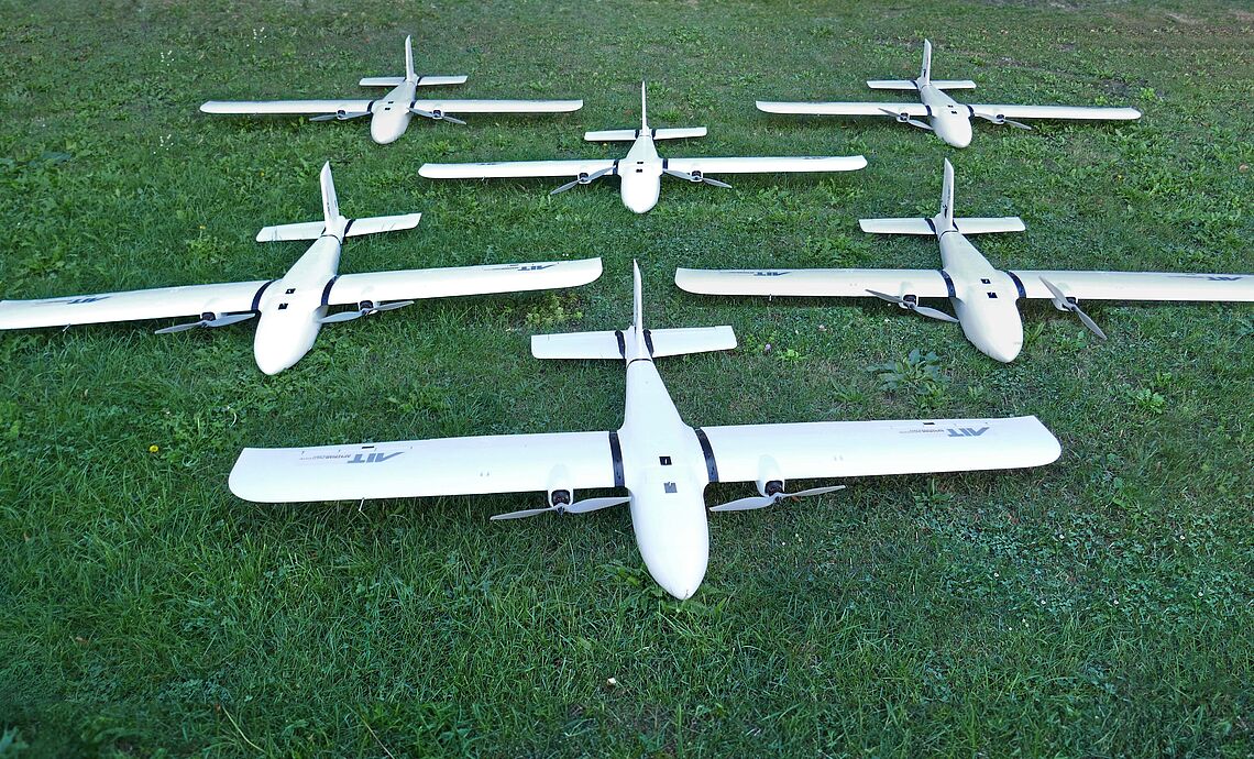 Several unmanned aerial vehicles equipped with sensors and the AIT logo lie on the meadow in front of the AIT Aerial Systems Lab