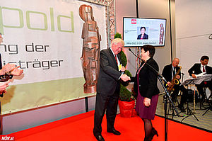 Angela Sessitsch betritt die Bühne auf der Wolfgang Knoll steht und ihr die Hand reicht, im Hintergrund der Bildschirm auf dem Angela Sessitsch als Preisträgerin verkündet wird