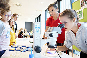 A group of children stands around a table with a microscope and samples in Petri dishes. A girl operates the microscope under the guidance of a scientist.