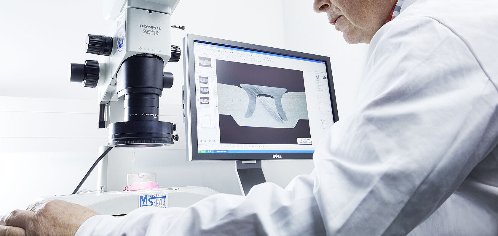Wissenschaftler untersucht Metallprobe mit Mikroskop vor einem Monitor