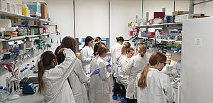 Ein Gang eines Labors in dem sich zwölf Schülerinnen und zwei Expertinnen aufhalten und arbeiten, alle tragen Laborhandschuhe und Labormäntel.
