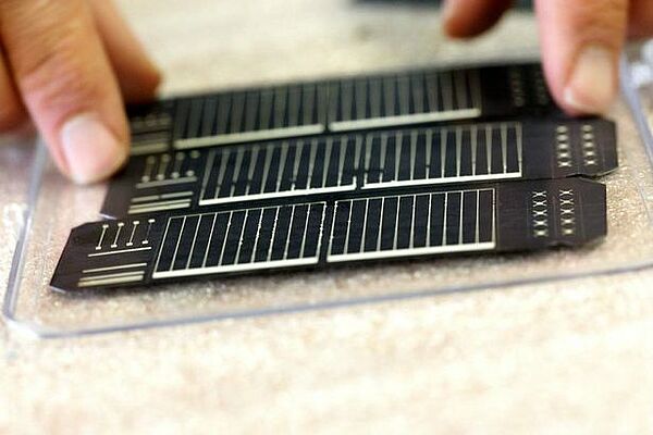 Zeigefinger einer Person berührt 3 kleine ultradünne Solarzellen, die auf einer Plexiglasunterlage liegen; copyright Sunplugged