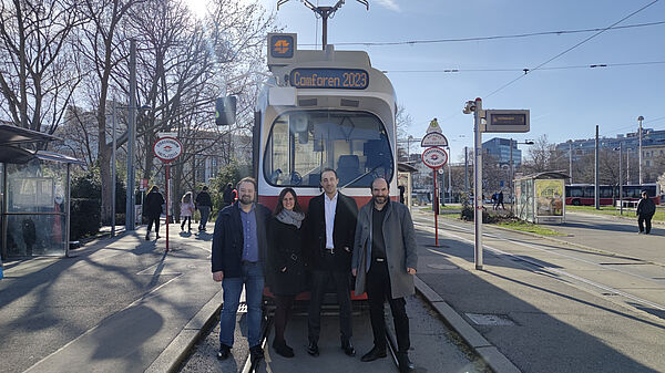 Foto der Organisatoren von ComForEn (Stefan Wilker, Carina Schöfl, Mark Stefan und Friederich Kupzog) vor einer Wiener Straßenbahn auf der ComForEn 2023 steht