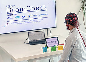 Mann sitzt vor einem Tablet und bunte Würfel liegen vor ihm, auf dem Kopf hat er ein Netz mit dem seine Gehirnwellen aufgezeichnet werden, an der Wand leuchtet ein Bildschirm mit dem Schriftzug "Braincheck"