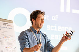 Walter Kuba präsentiert mit einem Molekül-Modell in der Hand vor seinem Poster.