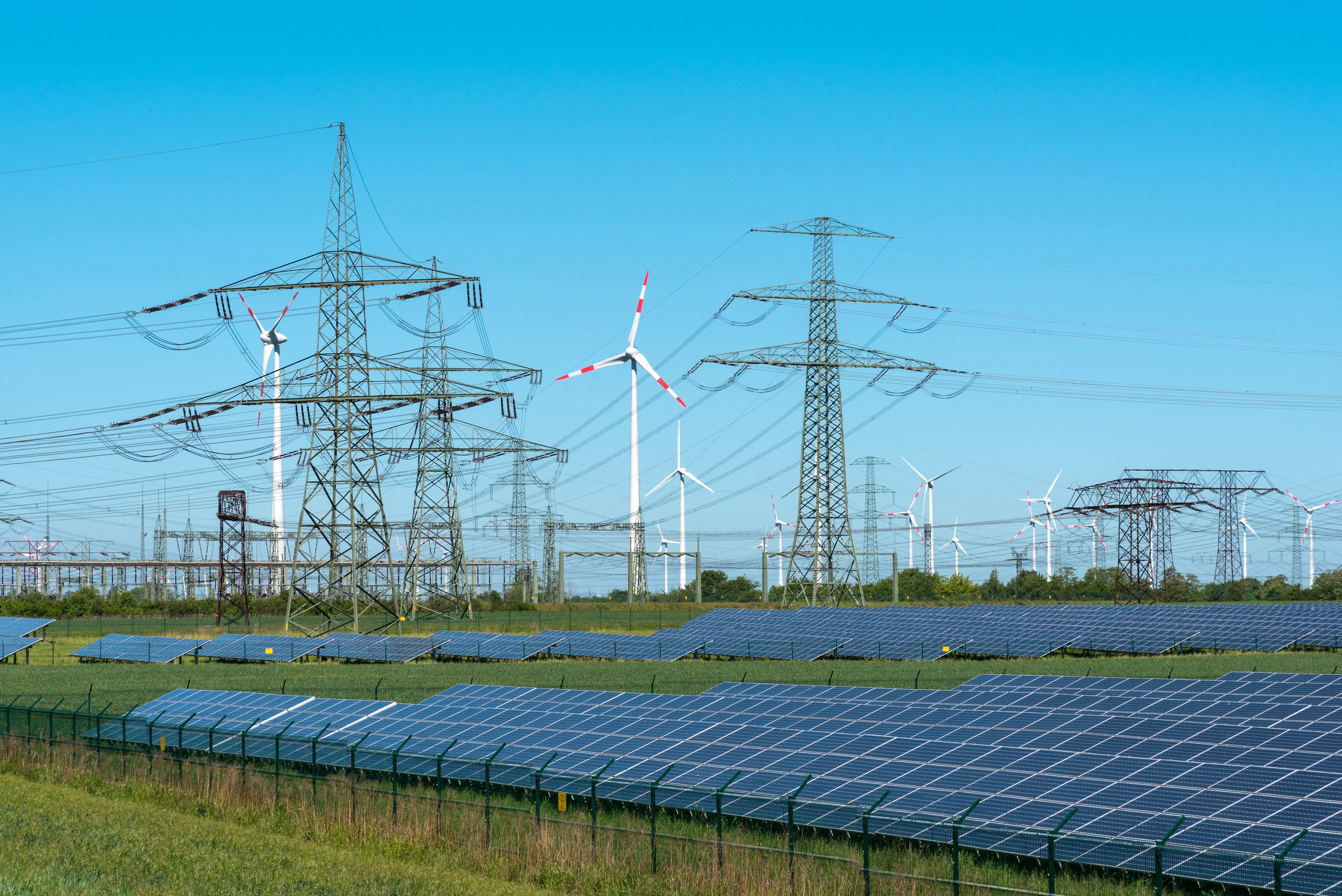 Symbolbild: Blauer Himmel und grüne Wiese großflächig mit Solaranlagen im Vordergrund. Weiter hinten sieht man Strommasten und Windräder.