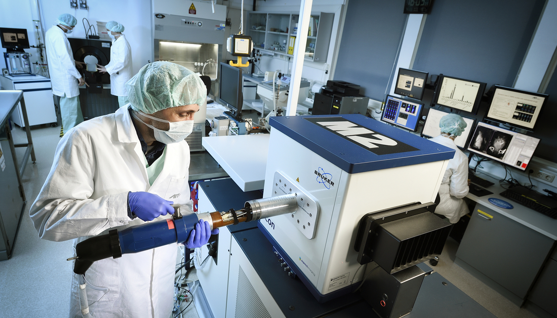 Labor mit arbeitenden Wissenschaftlern mit Haarnetzen und Schutzmasken die gerade einen Test vorbereiten indem eine Schiene in ein Testgerät eingeführt wird. Im Hintergrund weitere arbeitende Wissenschaftler.