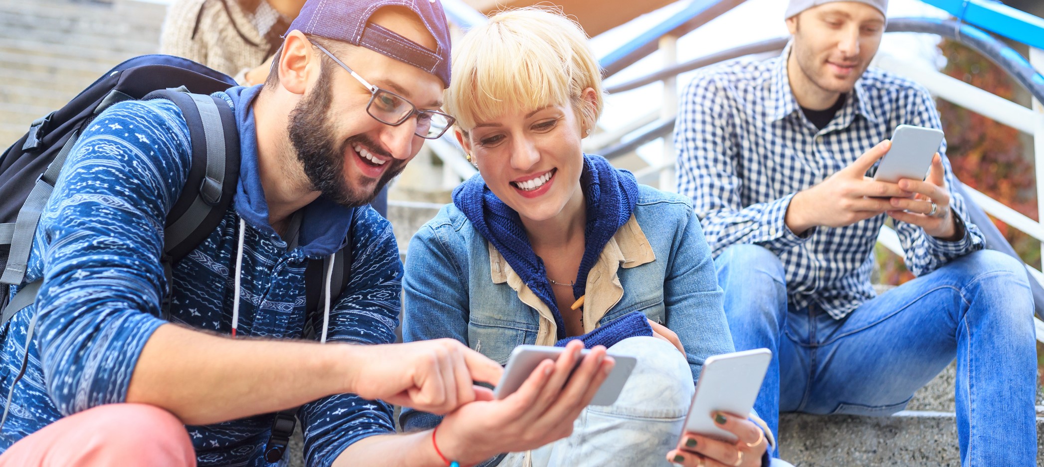 Man and woman sit on steps and look at smartphones. In the background there is another man.